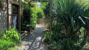 Um caminho no jardim leva a uma casa de madeira com pérgola, cercada por muita vegetação e diversas plantas.