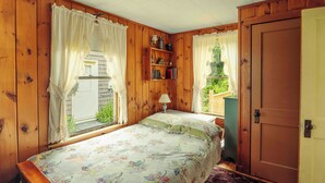 A bedroom with wooden walls, a bed with floral bedding, a nightstand, a lamp, a bookshelf, and a window with curtains. - Howard Family Cottage with Casco Bay Sunset Views! (Harpswell)