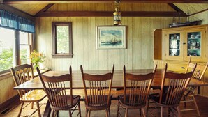 A wooden dining room with a large table and chairs, a window with a view, and a cabinet with glass doors. - Howard Family Cottage with Casco Bay Sunset Views! (Harpswell)