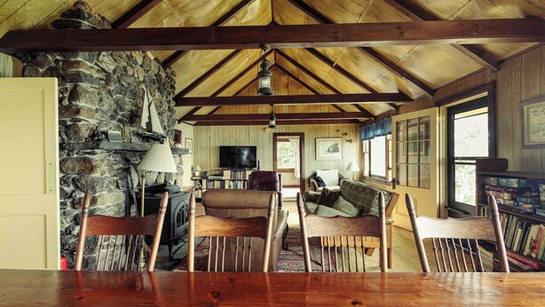 A rustic cabin interior with a stone wall, wooden ceiling beams, a fireplace, a television, and a cozy seating area. - Howard Family Cottage with Casco Bay Sunset Views! (Harpswell)