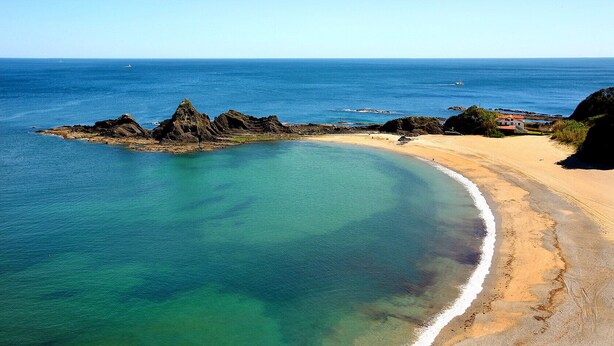 Paisatge costaner amb una platja sorrenca, aigües turqueses cristal·lines i formacions rocoses.