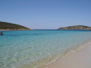 A beach with clear turquoise water, sandy shore, and a small hill with a structure on top.