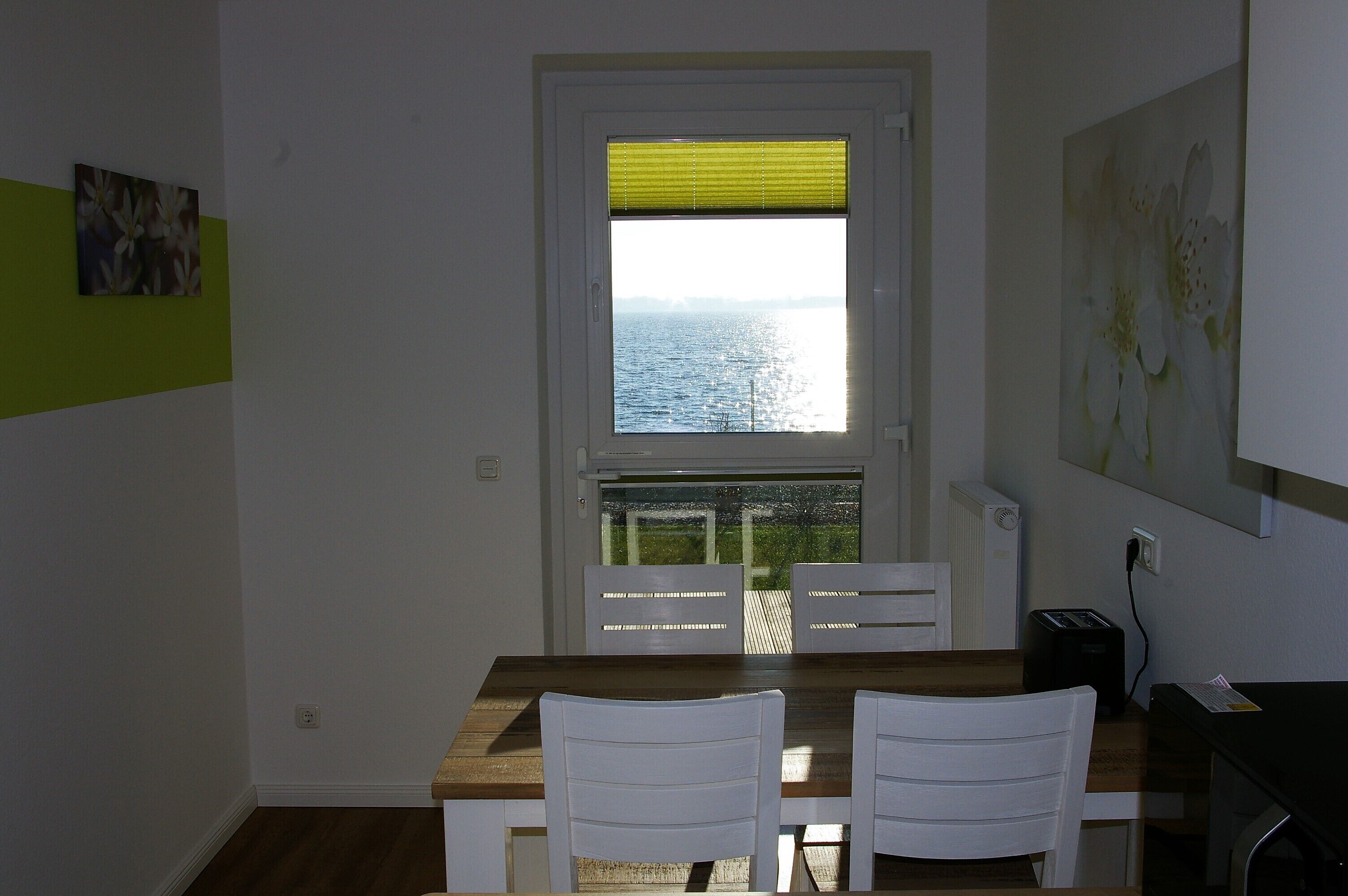 A dining area with a table and chairs, a window with a view of the sea, and a painting on the wall.
