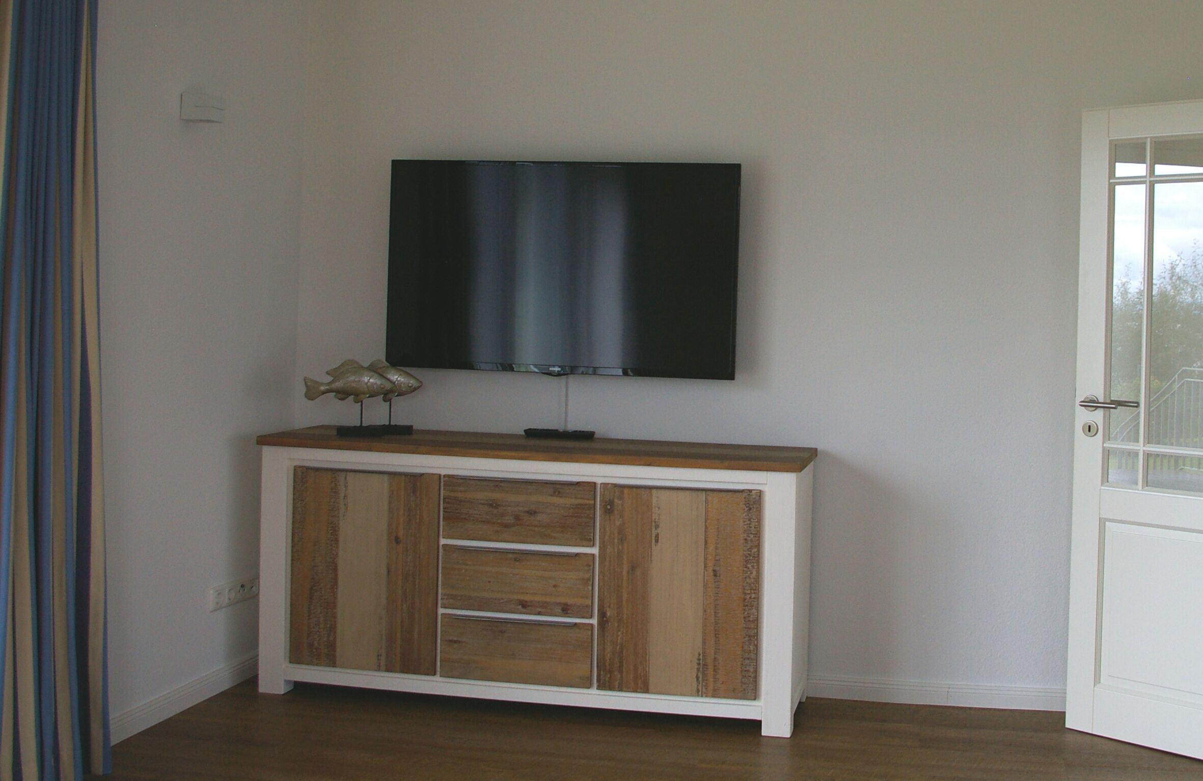 Une pièce avec un meuble en bois sous un téléviseur à écran plat, une porte avec un panneau de verre et un rideau sur le côté gauche.