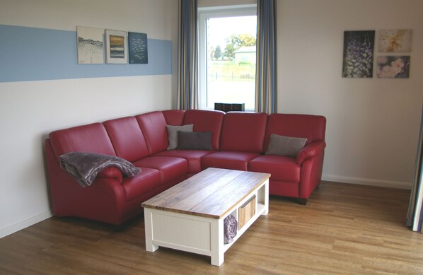 A red sectional sofa with a wooden coffee table in front, a window with curtains, and wall art. - Witt am See - Holidays at Wittensee (Klein Wittensee)
