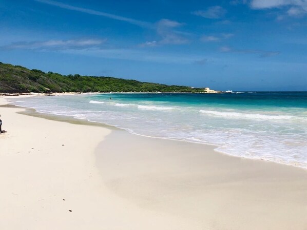 En strand med klart turkis vand, sandet bredde og en person stående til venstre.