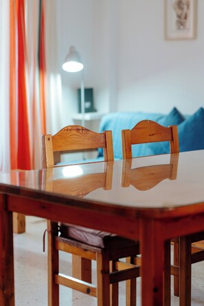 A wooden dining table with two chairs, a lamp in the background, and a bed with blue bedding.