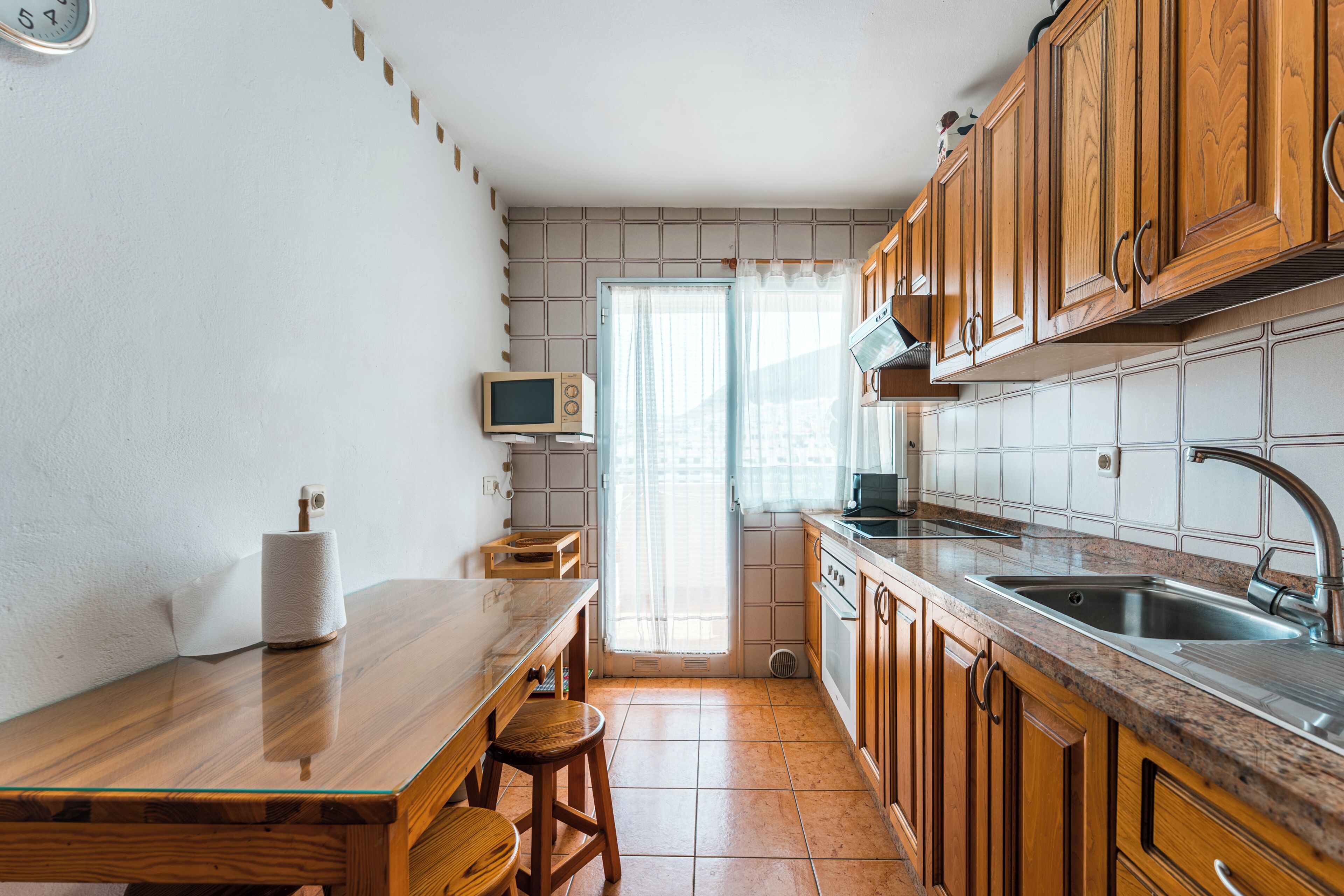 Una cocina con armarios de madera, un lavamanos de acero inoxidable, un microondas y una ventana con cortinas.