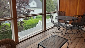 Un solárium con amplios ventanales, una terraza de madera, mobiliario metálico y vistas a una casa y follaje.