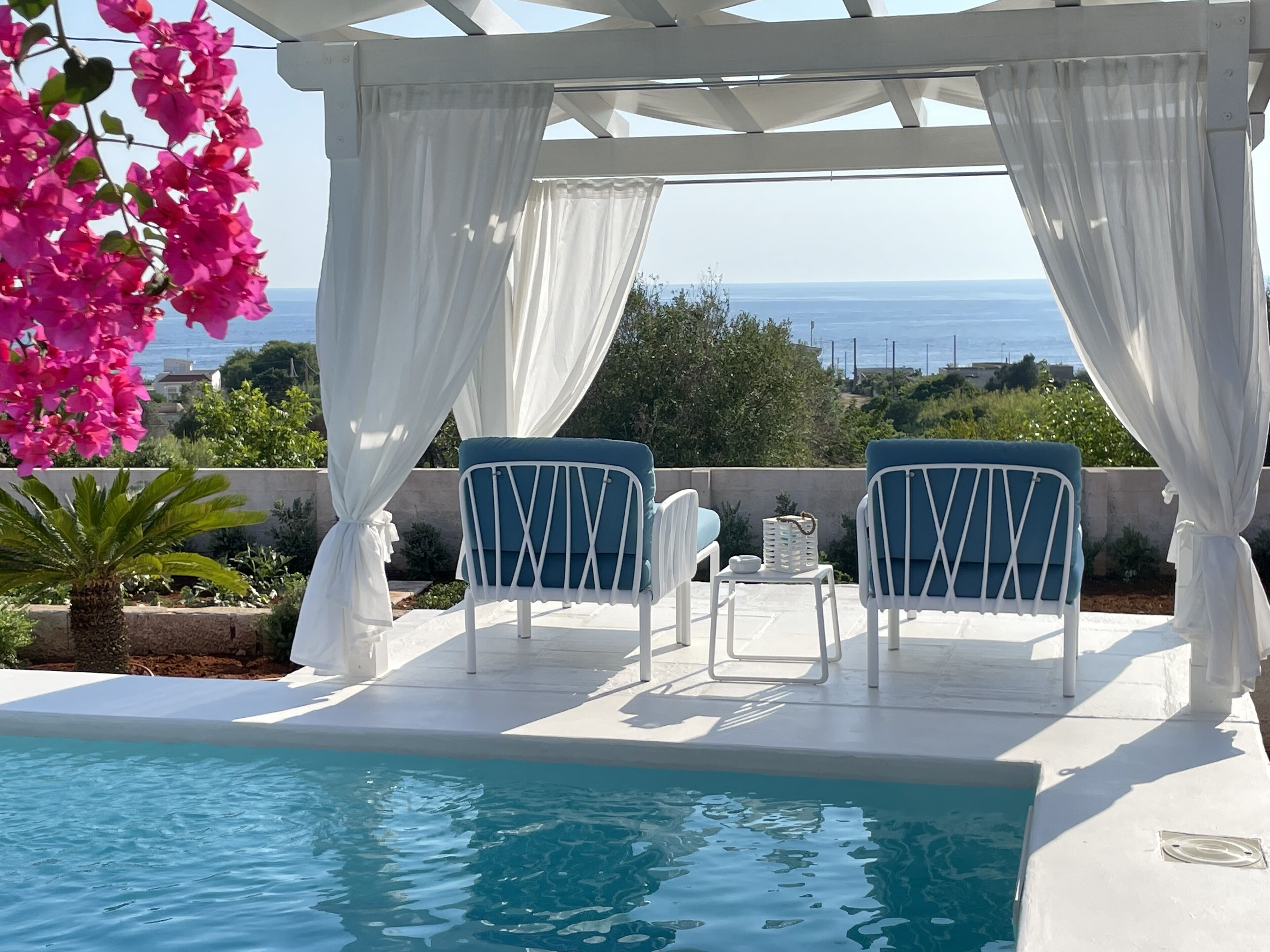 A poolside area with two blue cushioned chairs, white curtains, and a view of the sea.