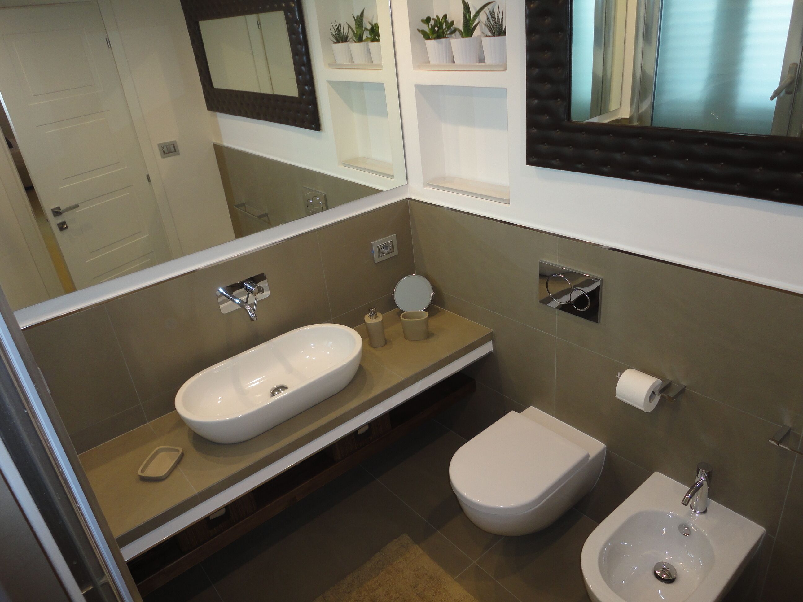 A modern bathroom with a large mirror, a white sink, a bidet, and a toilet. There are potted plants on a shelf above the mirror.