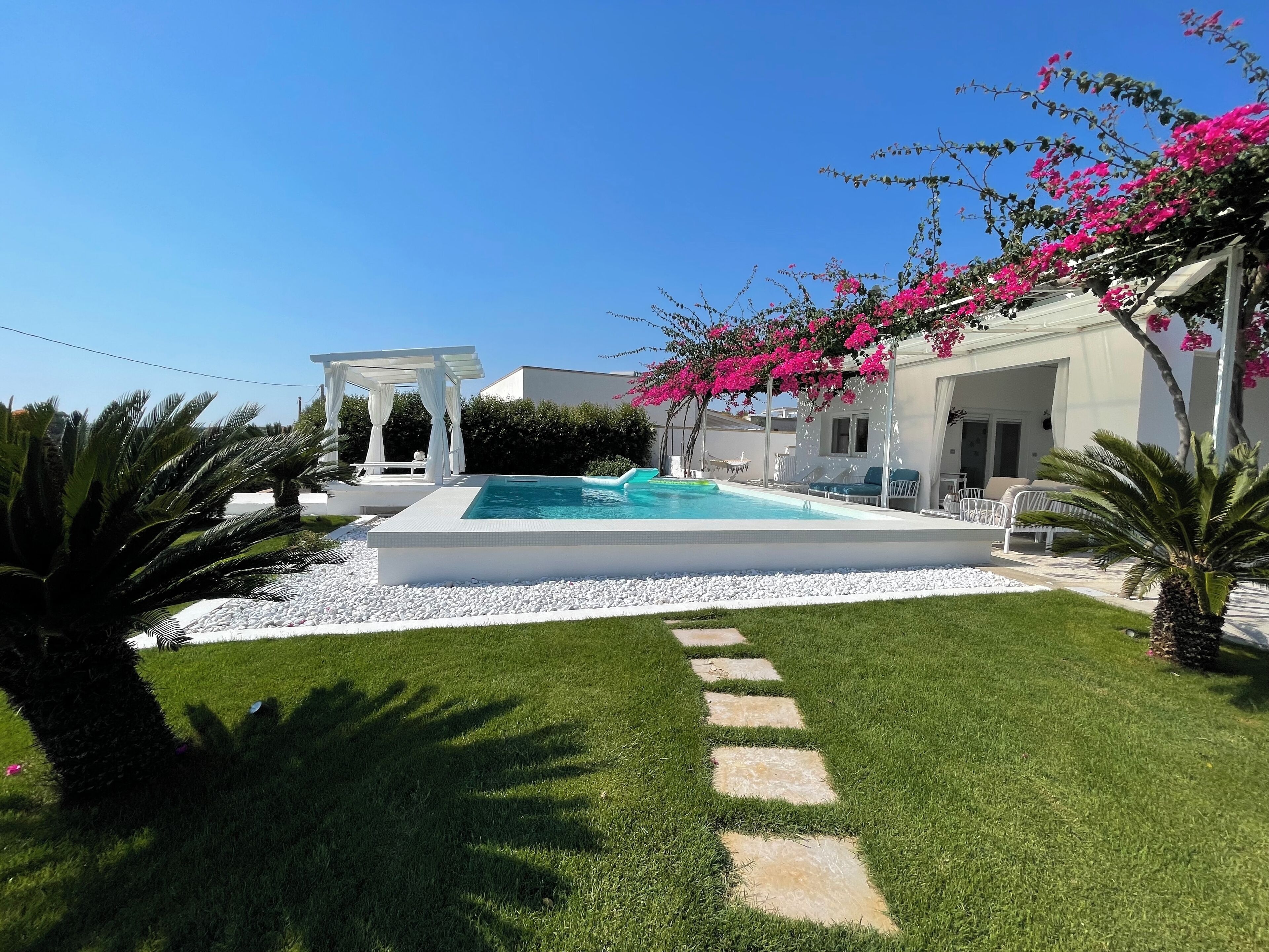 Ein Poolbereich mit weißer Pergola, Liegestühlen und blauem Himmel.