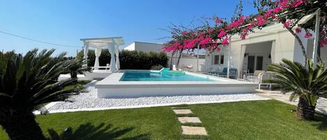Ein Poolbereich mit weißer Pergola, Liegestühlen und blauem Himmel.