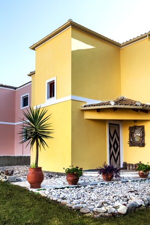 A yellow building with a tiled roof, a small window, and a decorative door. There are potted plants and a stone pathway in front.