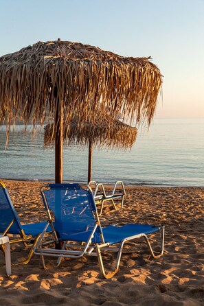 A beach with two blue lounge chairs and a straw umbrella.