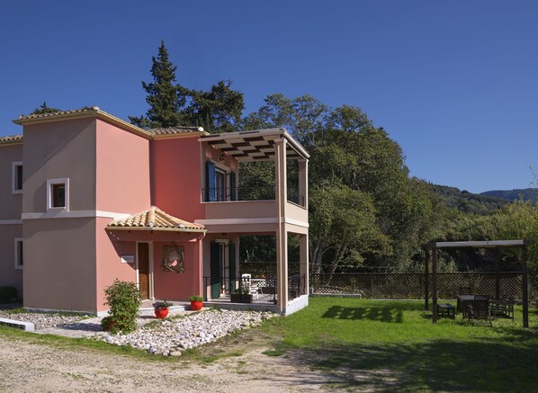 A two-story house with a balcony, a garden with potted plants, and a view of a forested hill.