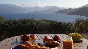 Een tafel met croissants, brood en een schaal druiven, met uitzicht op de kust en bergen op de achtergrond.