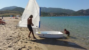 Una spiaggia con una barca a vela vicino alla riva, una persona che vi sta accanto e un'altra persona che si china in acqua.