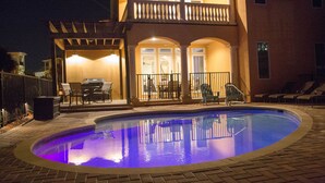 Una piscina con un patio perimetral, un área de estar al aire libre cubierta y un edificio de dos pisos con balcones.