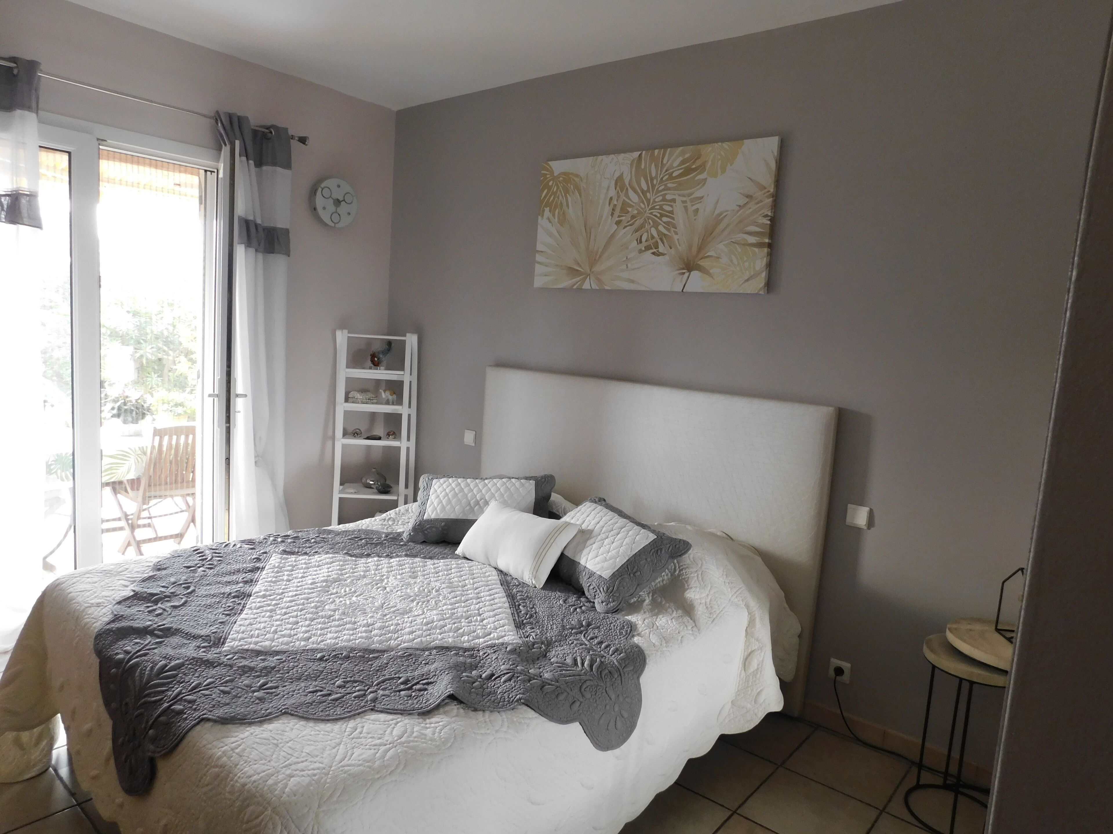 Un dormitorio con una cama grande, cabecera blanca, mesita de noche, un reloj de pared, estante con adornos y una ventana con cortinas.