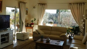 A living room with a beige sofa, a television, a coffee table, and a view of a garden through large windows. - Detached villa with good amenities, private pool, close to Aix en Provence (Coudoux)