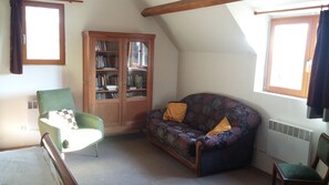 A cozy attic room with a green armchair, a patterned sofa, a wooden bookcase, and a window with curtains.