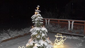 Ein mit Schnee bedeckter Weihnachtsbaum mit Lichtern, ein Schneemann und ein Schlitten in einer verschneiten Winterlandschaft.