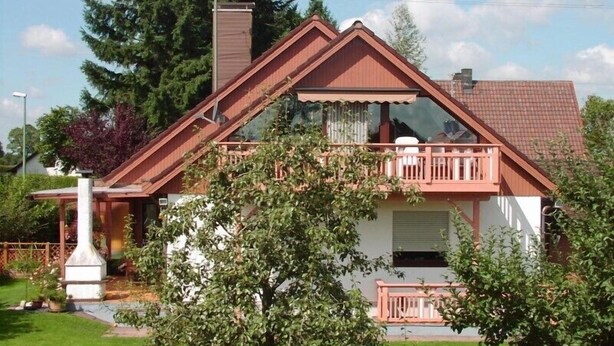 Ein zweistöckiges Haus mit rotem Dach, einem Balkon mit Geländer und einem Schornstein.