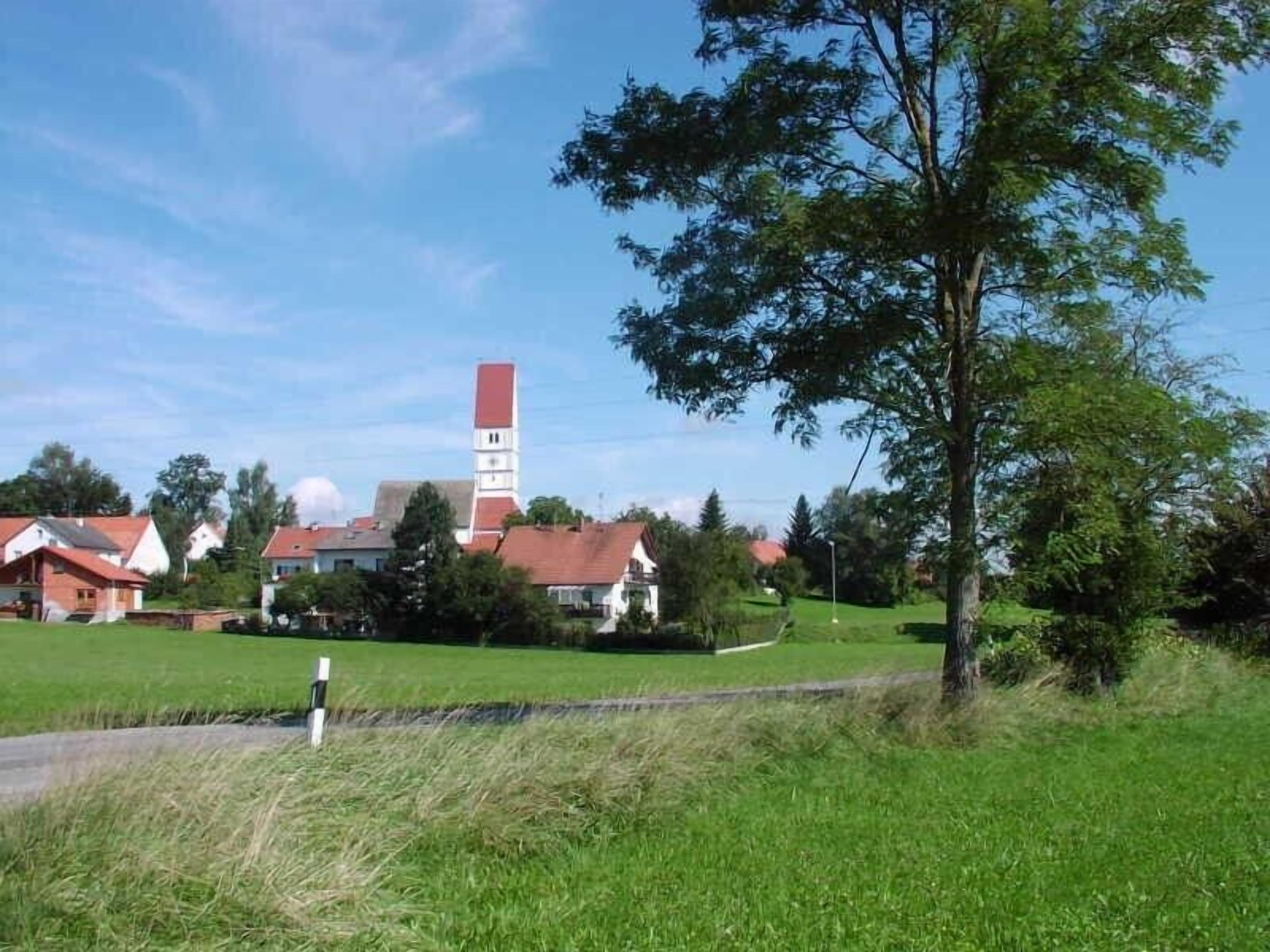 Un paysage rural avec une église dotée d’un haut clocher, entourée de maisons et de verdure.