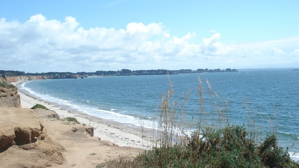 Paesaggio costiero con spiaggia sabbiosa, cielo azzurro limpido e forme del territorio in lontananza.
