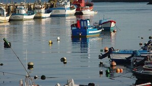 Ein Hafen mit mehreren Schiffen, ein Leuchtturm im Hintergrund und eine ruhige Wasserfläche.