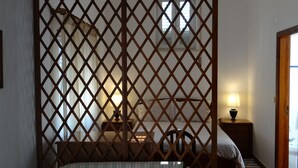 Ein Zimmer mit einem Holzgitterteiler, einem Bett mit floralem Überwurf, einem Nachttisch mit Lampe und Blick ins Freie durch ein Fenster.