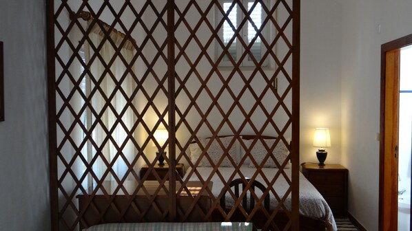 A room with a wooden lattice partition, a bed with a floral bedspread, a bedside table with a lamp, and a view of the outside through a window.