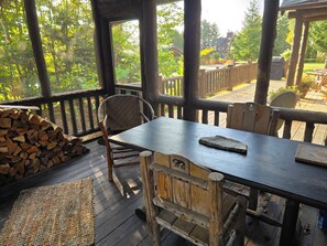 A wooden deck with a table and chairs, a firewood stack, and a view of a forested area.