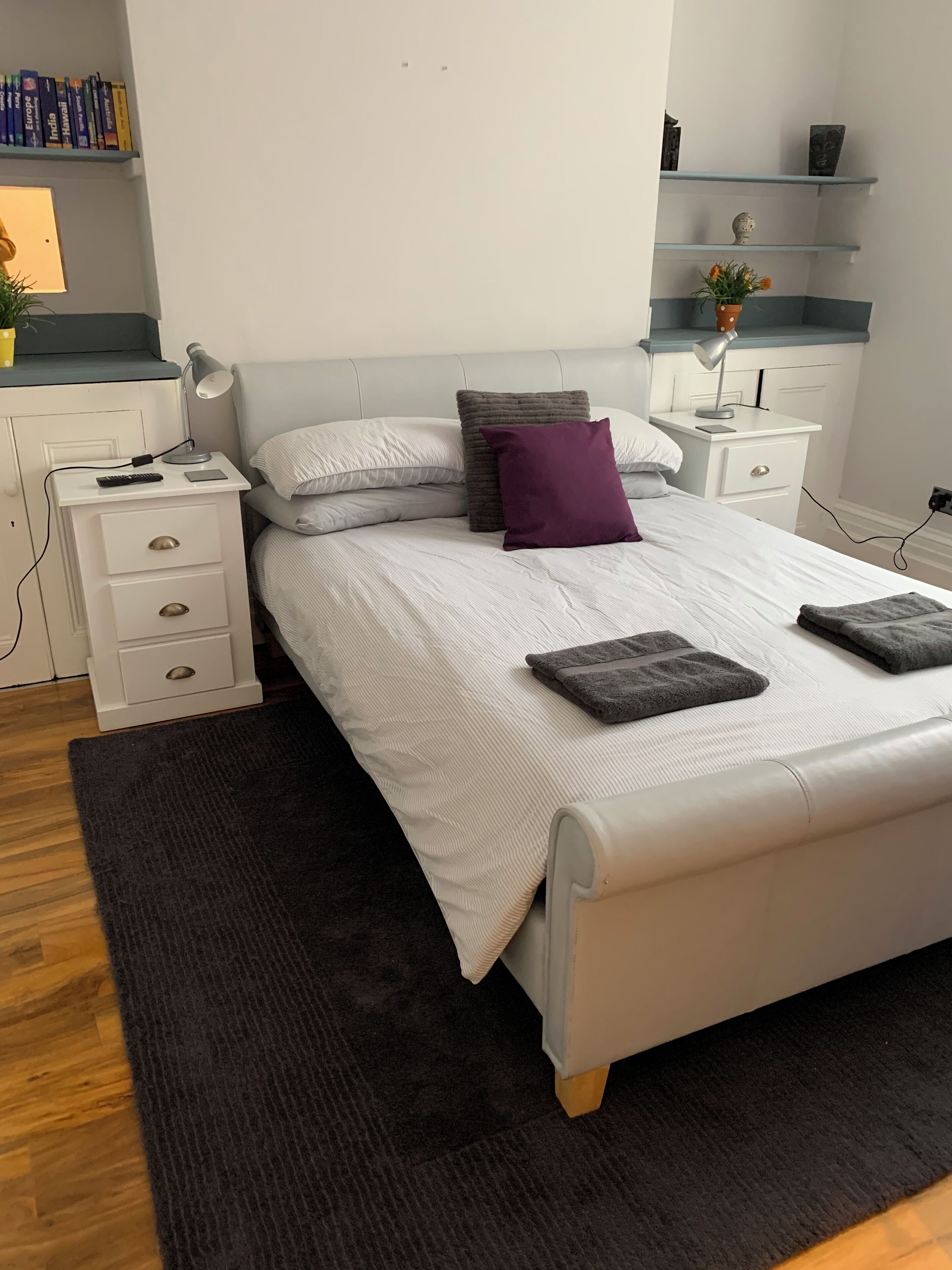 Een netjes ingerichte slaapkamer met een bed, nachtkastjes, een lamp, een plank met boeken en een kleine plant.
