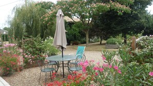 A patio area with a table and chairs, surrounded by various plants and flowers. - Quiet house between countryside and sea surrounded by green setting (Rouillé)