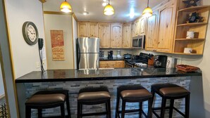 A kitchen with wooden cabinets, a stainless steel refrigerator, a microwave, and a granite countertop. There are three bar stools and a clock on the wall. - Luxury Condo w/ FREE WiFi, Parking, Heated Pool, Hot Tubs, Skier Shuttles (Avon)