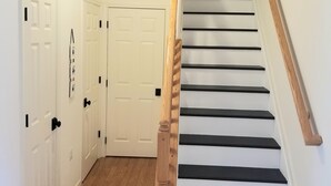 Una escalera con barandillas de madera y peldaños negros, que lleva a un rellano con puertas blancas y una lámpara en el techo.