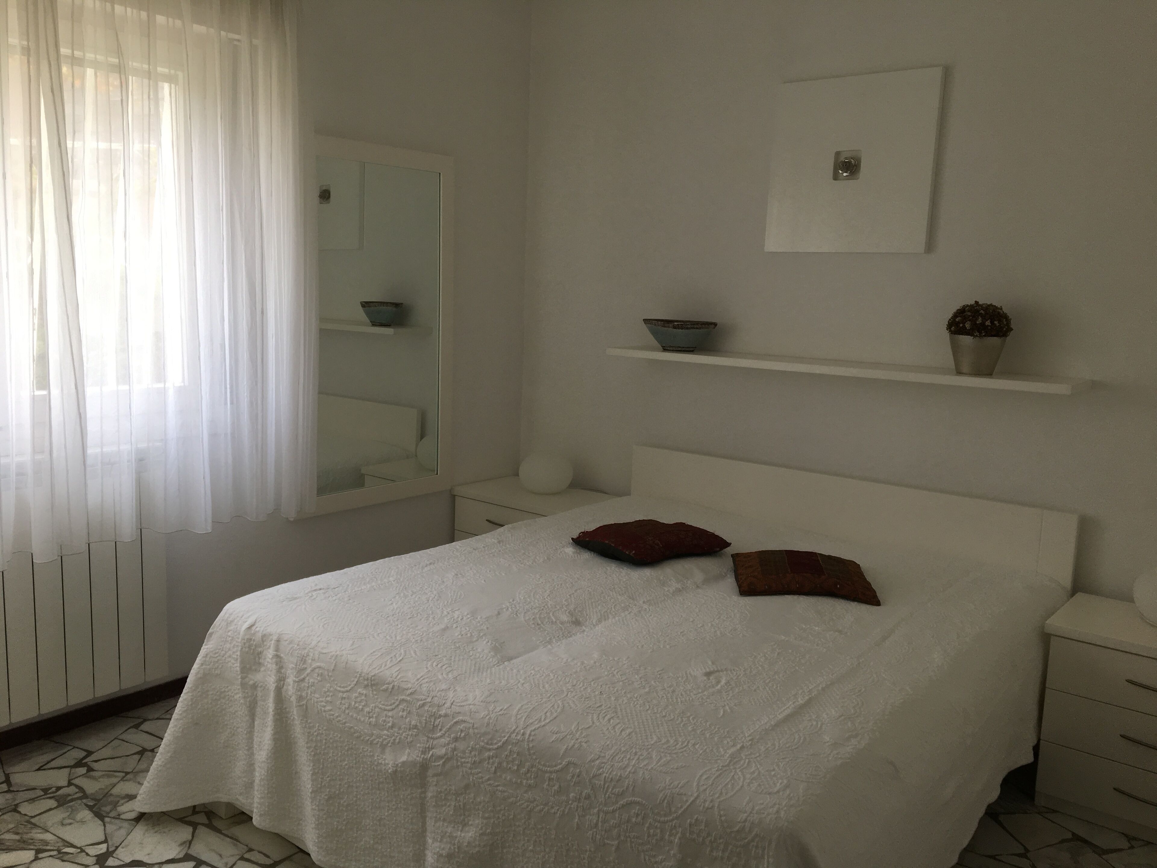 A bedroom with a bed, a nightstand, a mirror, a shelf with decorative items, and a window with sheer curtains.