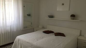 A bedroom with a bed, a nightstand, a mirror, a shelf with decorative items, and a window with sheer curtains.