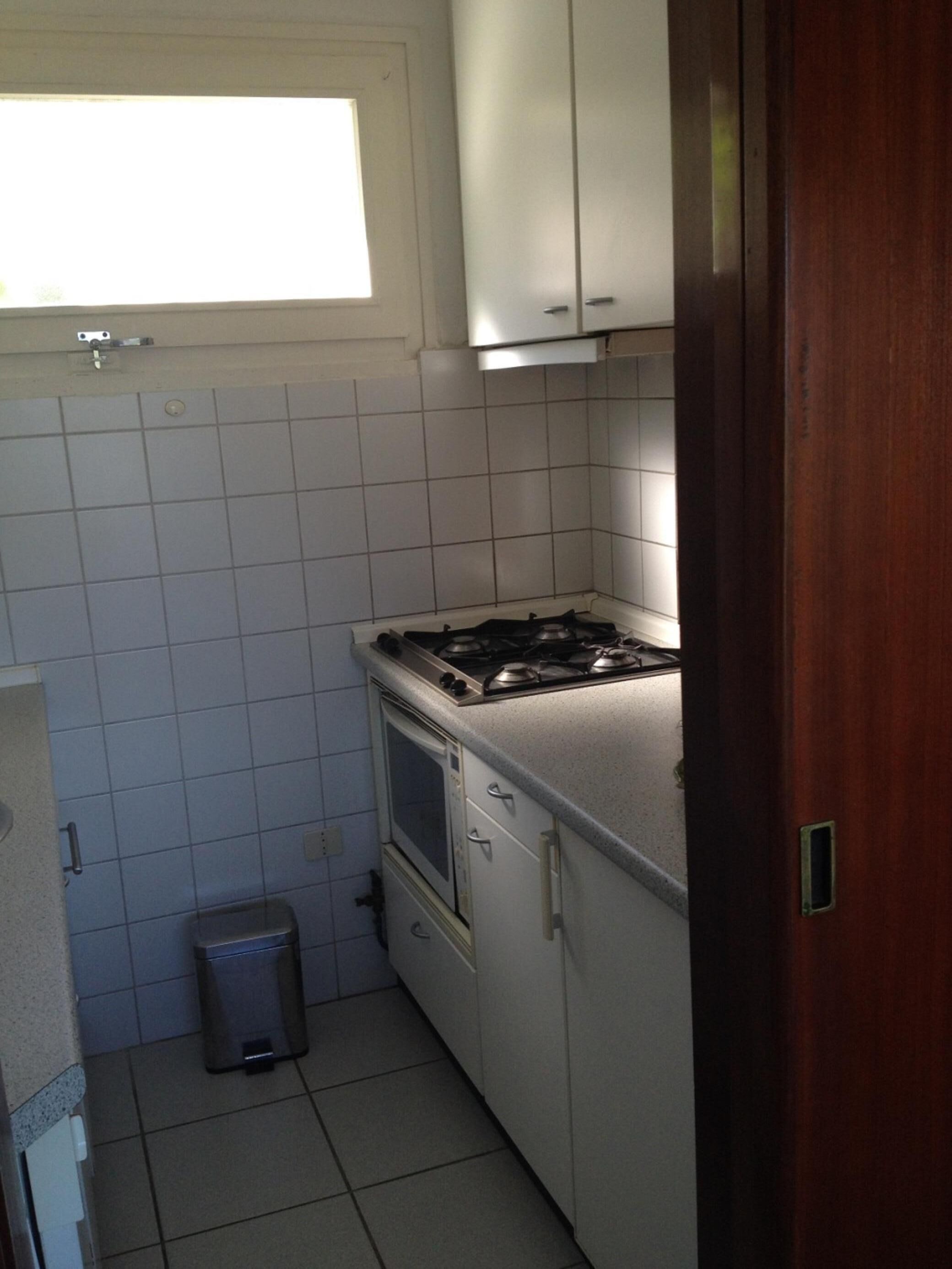 Une cuisine de taille réduite, équipée d’une cuisinière à gaz, d’un four et de meubles blancs. Il y a une fenêtre au-dessus de l’évier et une poubelle à côté du four.