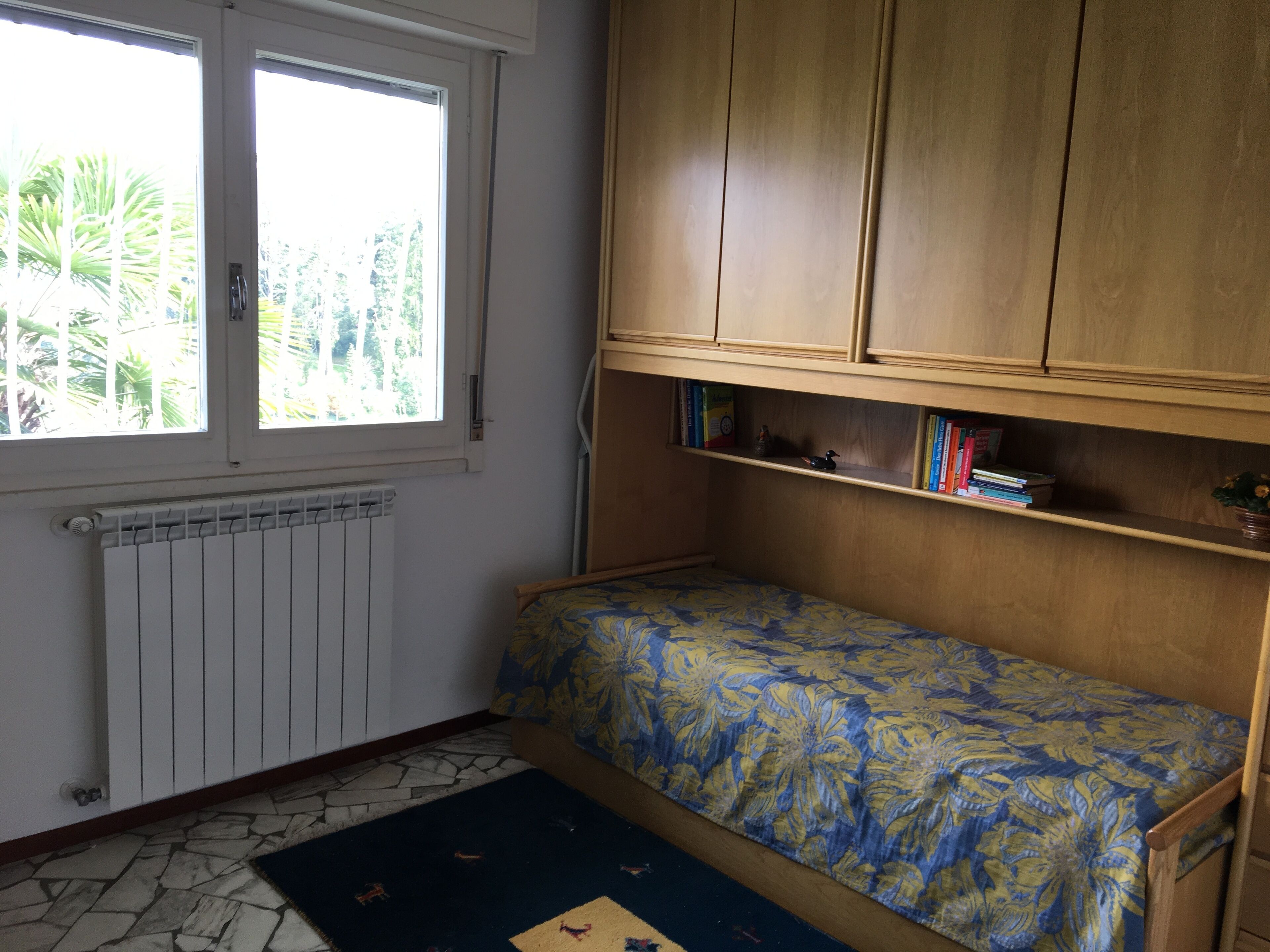 Een slaapkamer met een enkel bed, een radiator, een raam met uitzicht op groen en een boekenkast met boeken en een klein beeldje.