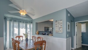 Un coin repas avec une table et des chaises en bois, un ventilateur de plafond et une vue sur la cuisine.