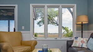 Un salon avec un fauteuil en cuir beige, un canapé marron, une table basse sur laquelle se trouvent une tasse et une télécommande, et une fenêtre donnant sur des arbres et des champs.