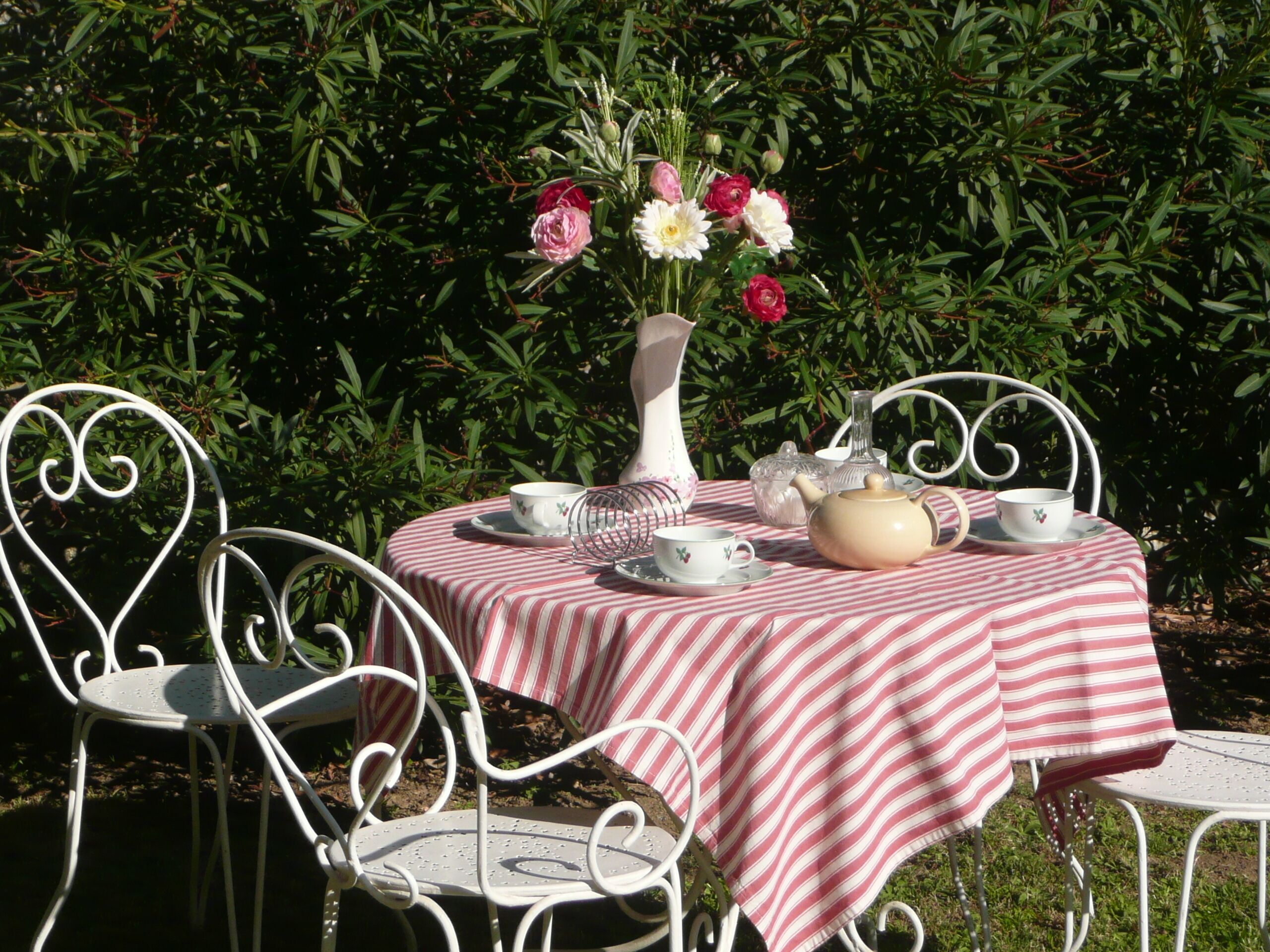 Une table dressée à l’extérieur, recouverte d’une nappe à rayures, entourée de chaises en métal blanc, et agrémentée d’un vase rempli de fleurs.