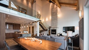 A spacious living area with a wooden dining table, grey sofa, and a stone fireplace. - TRUE ski in/out. ski_bike_hike to BOTH Gondolas-Pvt HotTub VIEWS-walk everywhere (Whistler)