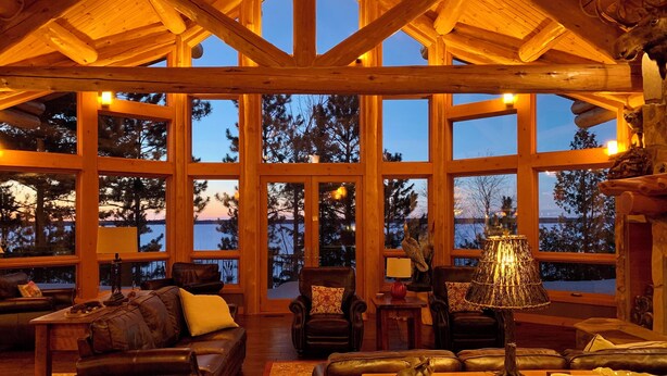 A spacious living room with large windows, wooden beams, leather sofas, and a fireplace. - Private, Exclusive Lodge on Woman Lake with Beautiful Views (Longville)