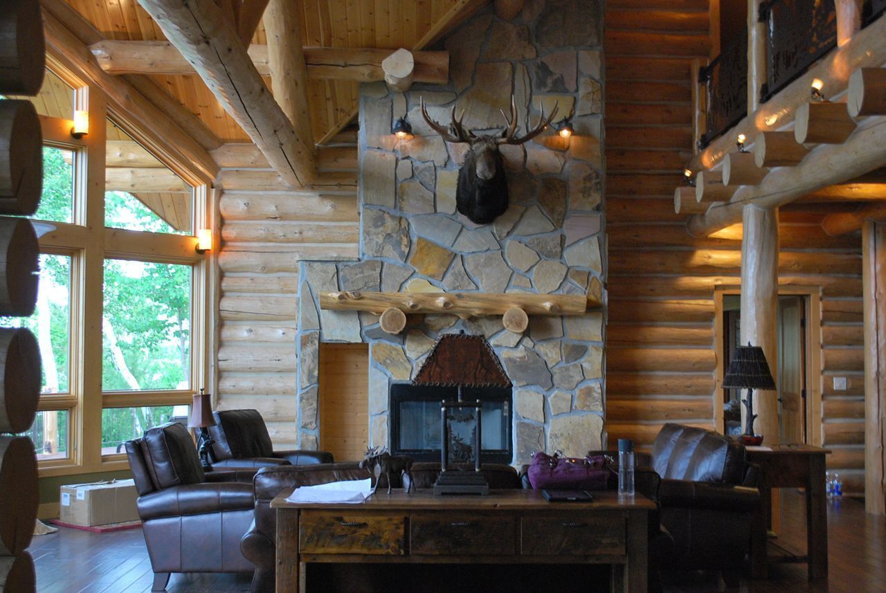 A log cabin interior with a stone fireplace, leather furniture, and a mounted deer head.