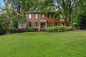 A two-story brick house with a front porch, surrounded by a well-maintained lawn and mature trees.