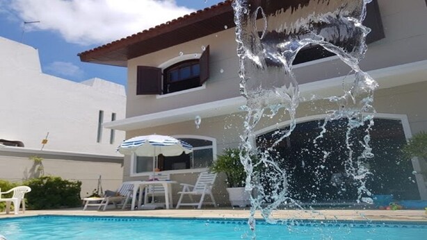 A swimming pool with water splashing, a covered patio with lounge chairs, and a building with a brown roof and windows. - Guarujá, Pernambuco Beach, a 3-minute walk from the beach. A paradise on the beach! (Balneario Praia do Pernambuco)